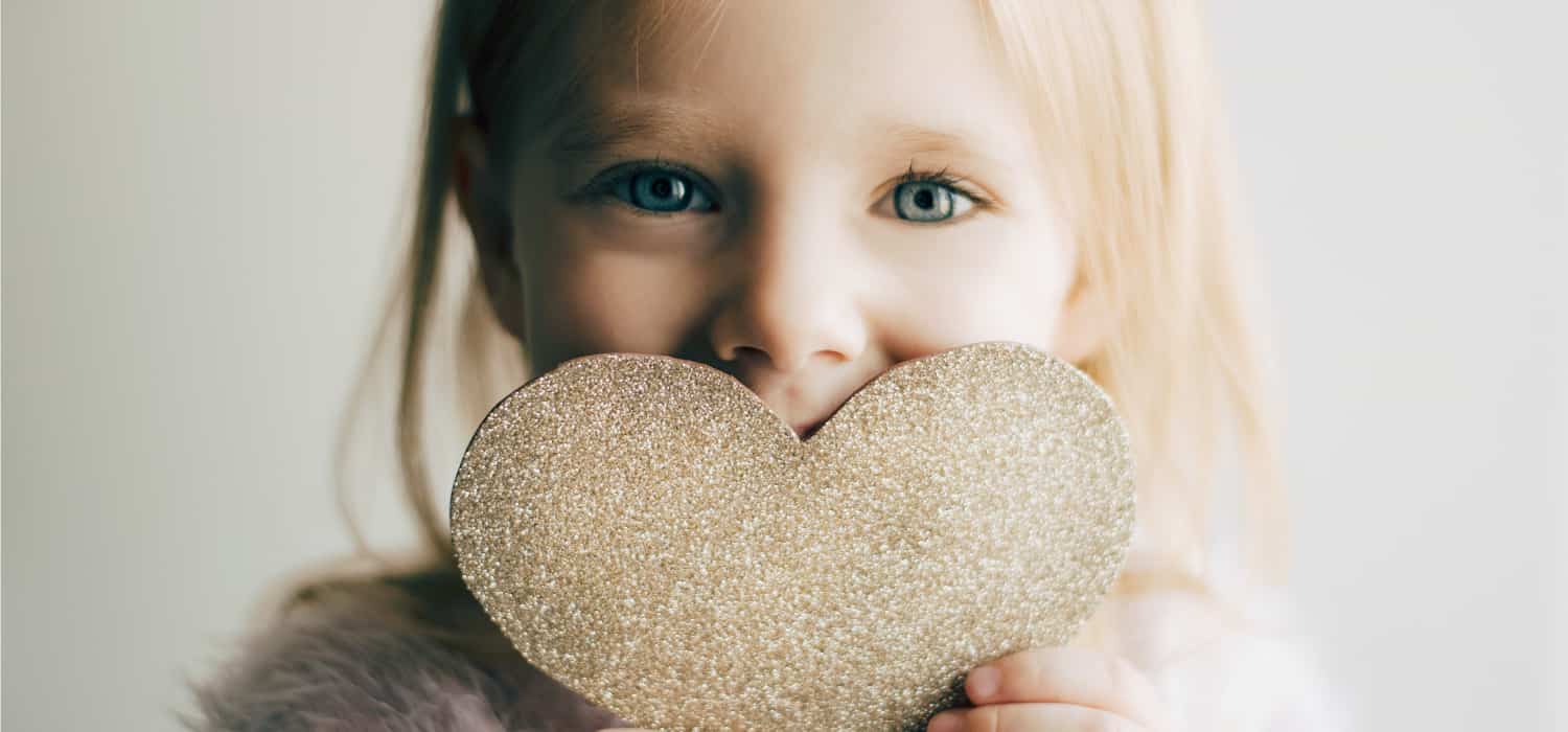 Girl with blonde hair holding a glitter heart in front of her mouth